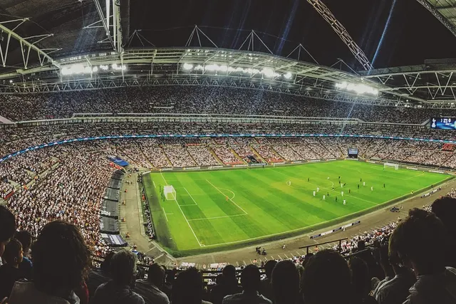 Panoramic view of a packed floodlit stadium during a night soccer match, with fans watching players on the pitch