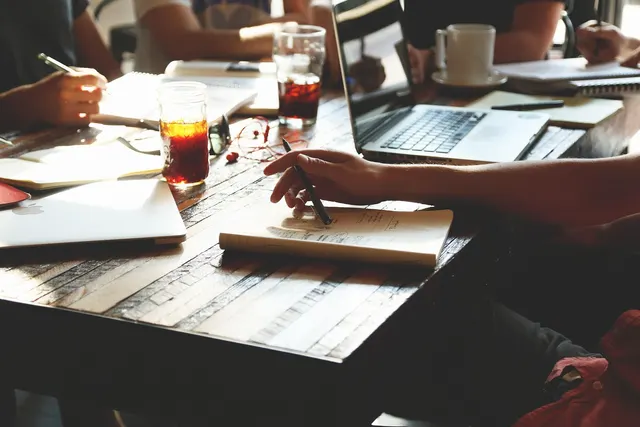 Team working at a rustic wooden table with laptops, notebooks, pens, iced tea glasses and a coffee mug