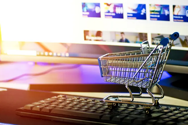 Miniature metal shopping cart on a black keyboard in front of a monitor showing a blurred online store page