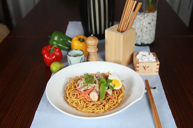 Bowl of noodles with egg and vegetables beside chopsticks and peppers