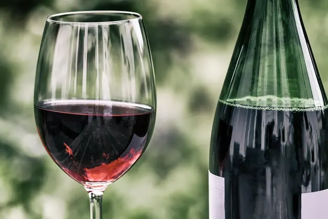 Glass of red wine beside a dark green wine bottle with a blank white label against a blurred green background