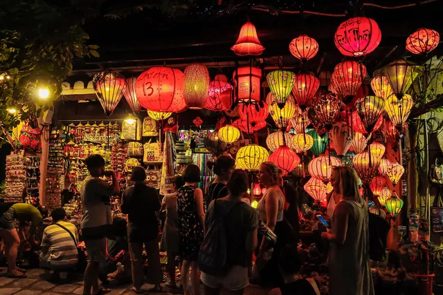 Tourists browsing a night market stall hung with glowing red and yellow silk lanterns, one checking a phone