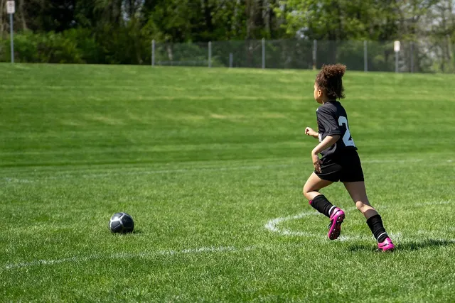 Young soccer player in a black team jersey and pink cleats running across a grass field toward the ball