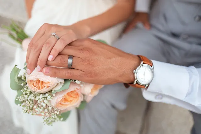 Couple's hands with a diamond engagement ring and wedding bands resting on a bouquet of peach roses
