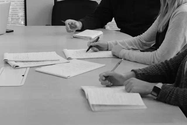 Black and white photo of three people taking handwritten notes on notepads at an office conference table