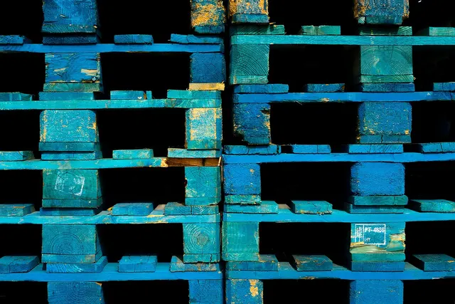 Close-up of stacked blue wooden shipping pallets with weathered paint, shown end-on against a dark background