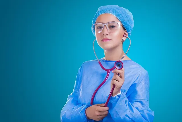 Healthcare worker in blue scrubs, surgical cap and safety glasses holding a pink stethoscope against a teal background