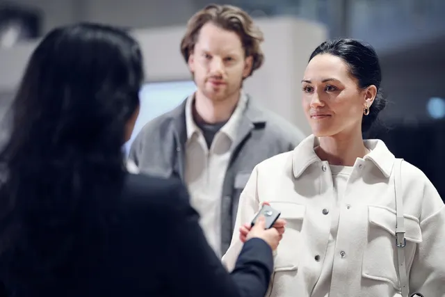 Dealership salesperson hands a car key fob to a smiling woman in a white jacket as a man looks on in a showroom