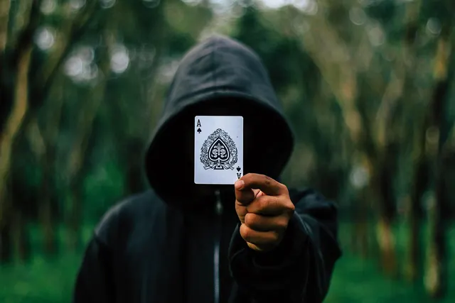 Person in a black hoodie hides their face behind an ace of spades playing card in a blurred green forest