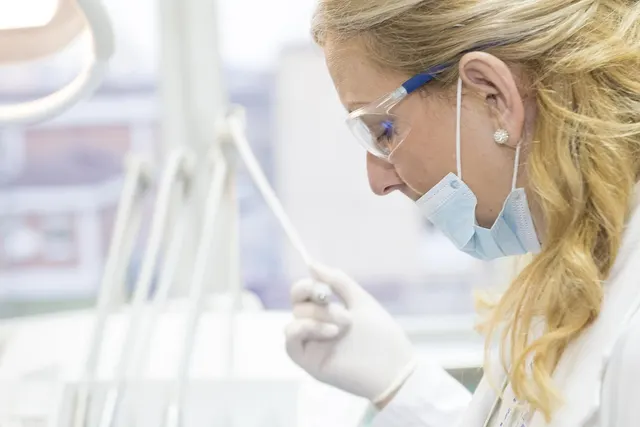 Dentist in safety glasses, face mask and white gloves holding a dental instrument beside a dental chair light