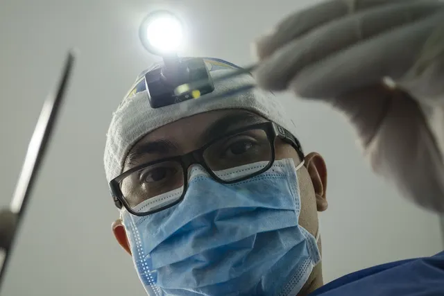 Dentist in glasses, surgical mask and headlamp leans over a patient holding dental instruments, seen from the chair
