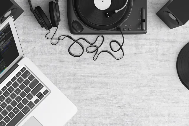 Overhead view of a laptop, headphones, turntable, vinyl records and speakers on a pale wooden desk