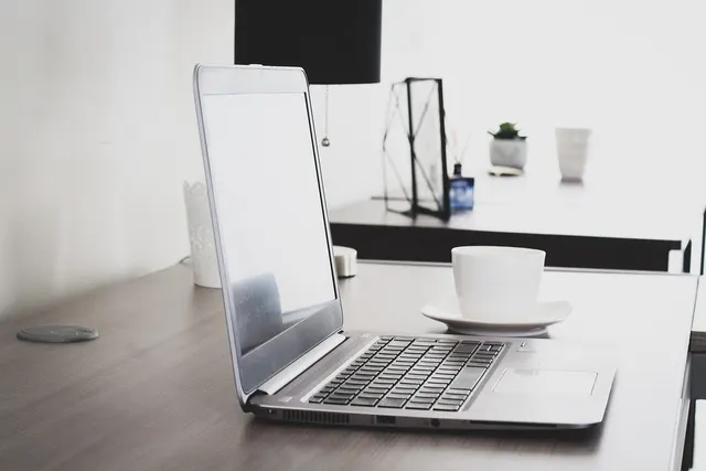 Silver laptop with a blank screen beside a white coffee cup and saucer on a wooden desk in a minimalist office