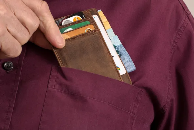 Hand pulling a brown leather wallet holding payment cards and cash from the pocket of a maroon button-up shirt