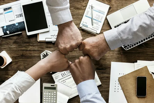 Four business people fist bump over a wooden desk with an invoice clipboard, calculator, tablet, laptop and paperwork