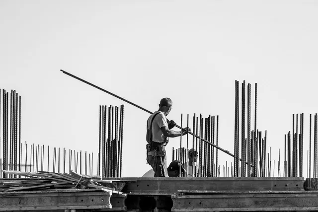 Black-and-white photo of a construction worker carrying a long steel rebar across a building site with vertical rebar