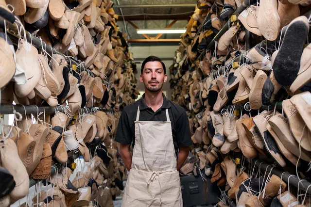 Craftsman in a canvas apron standing in a narrow aisle between tall racks of hanging wooden shoe lasts in a workshop
