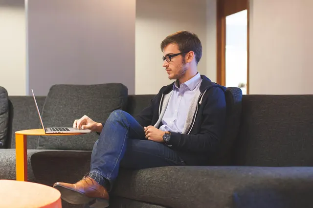 Young man in glasses and hoodie working on a laptop on a small orange table while seated on a gray sofa