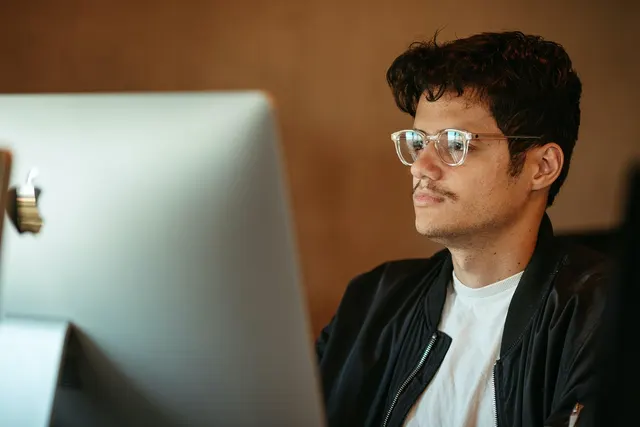 Young man with clear glasses and dark jacket intently reading a large silver desktop computer monitor