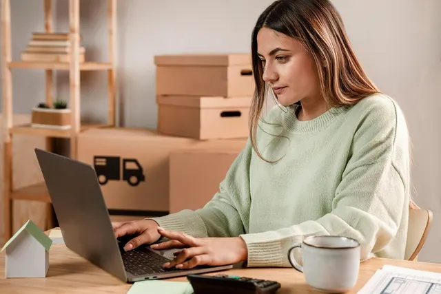 Woman in a green sweater typing on a laptop at a desk with a calculator and mug, shipping boxes stacked behind her