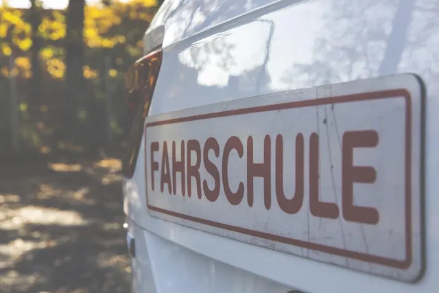 Close-up of a red and white 'Fahrschule' driving school sign on the back of a white car, autumn trees behind