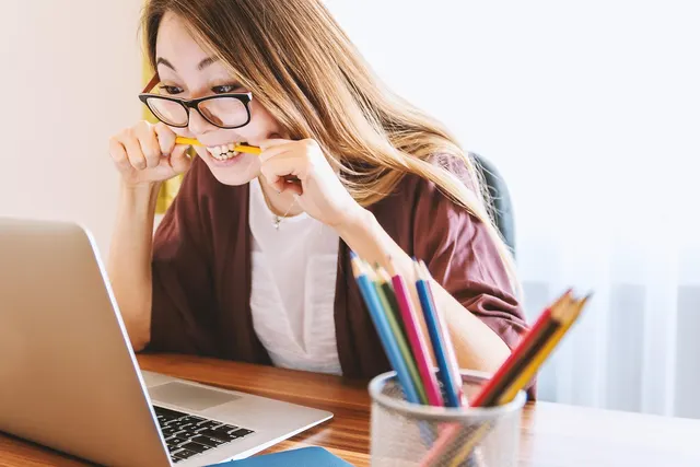 Frustrated woman in glasses biting a pencil while staring at her laptop at a desk with a cup of colored pencils