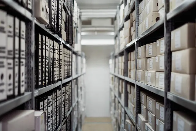 Narrow archive aisle with metal shelves holding black binders on the left and labeled cardboard boxes on the right