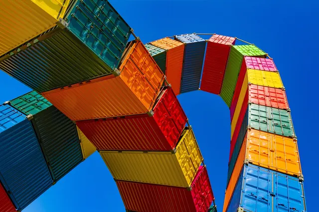 Colorful stacked shipping containers forming a tall arch against a bright blue sky, viewed from below