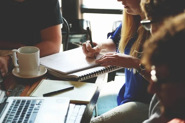 Team at a wooden cafe table with laptops, coffee and notebooks as a woman in a blue shirt writes notes
