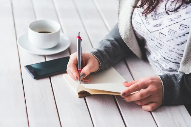 Woman writing in a small notebook at a white wooden table beside a smartphone and a cup of black coffee