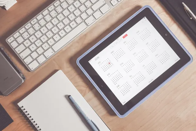 Tablet showing a yearly calendar on a wooden desk beside a white keyboard, smartphone, spiral notebook and pen