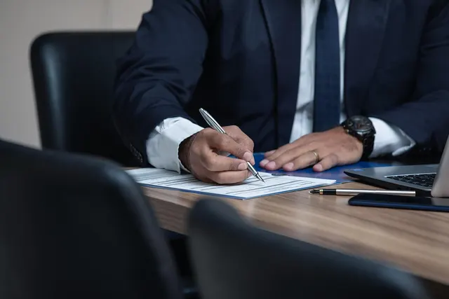 Professional in a navy suit signing a document at a conference table beside a laptop, fountain pen and phone
