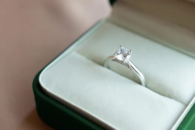 Solitaire diamond engagement ring with a pavé band resting in an open green ring box with cream velvet lining