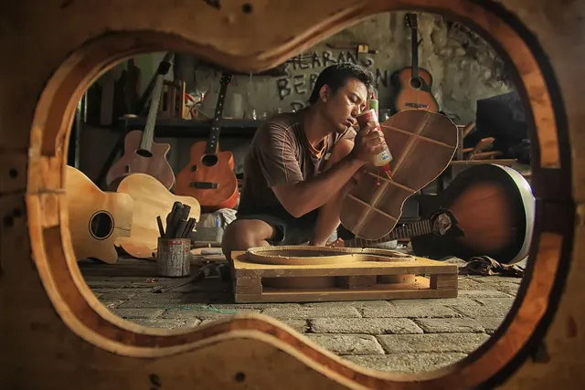 Luthier gluing braces onto a guitar back in a workshop full of handmade guitars, framed by a guitar body mold