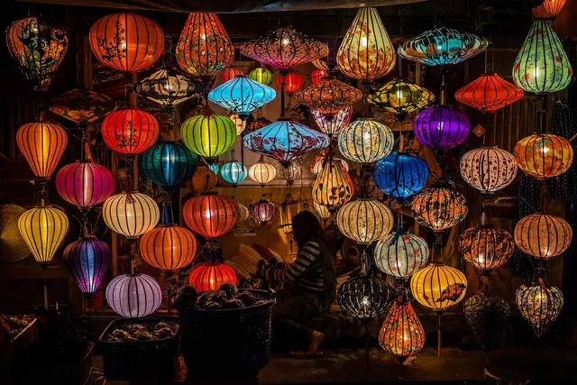 Vendor seated among glowing handmade silk lanterns in many colors hanging in a small craft shop at night