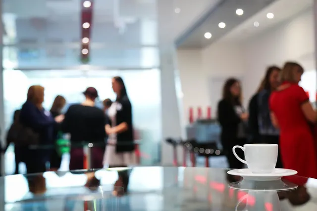 White coffee cup on a glossy table with blurred businesspeople talking at a bright office networking event