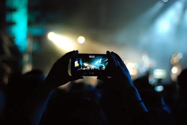 Silhouetted hands hold a smartphone filming a concert stage lit by spotlights and haze above a dark crowd