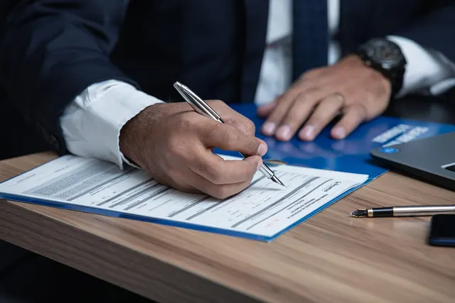 Man in a dark suit signing a printed form on a blue folder at a wooden desk, with a fountain pen and laptop nearby