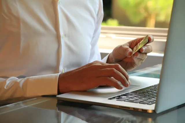 Person in a white shirt holding a gold credit card while using a laptop trackpad at a glass desk by a window