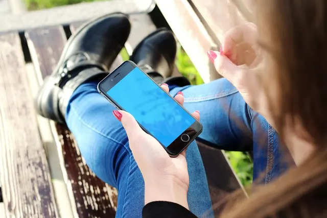 Woman in jeans and black boots sitting on a wooden bench, holding a smartphone with a blank blue screen