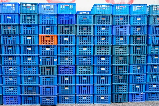 Wall of stacked blue plastic shipping crates with small labels, one bright orange crate standing out among them