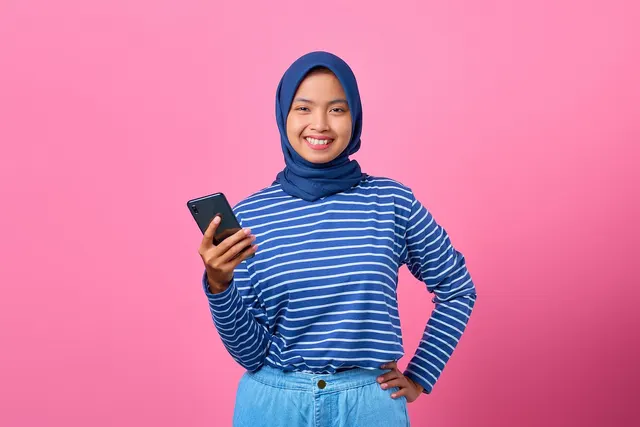 Smiling young woman in a blue hijab and striped shirt holding a smartphone against a pink background
