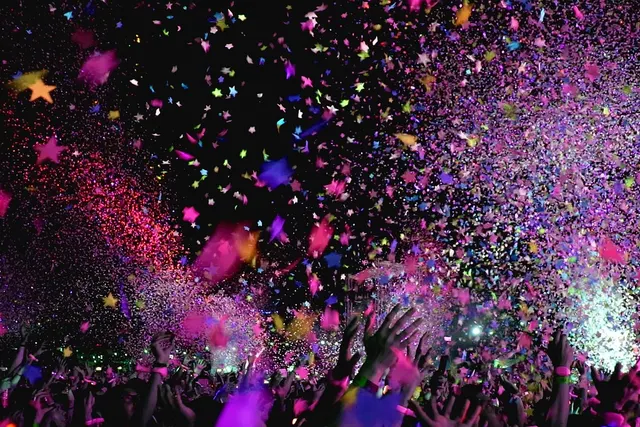 Festival crowd with raised hands under a shower of pink, purple and star-shaped confetti lit by stage lights