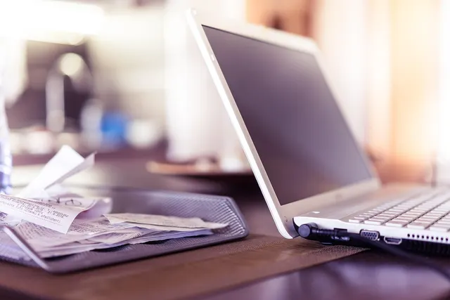 Silver laptop on a wooden desk beside a mesh tray piled with paper receipts, ready for tax and bookkeeping