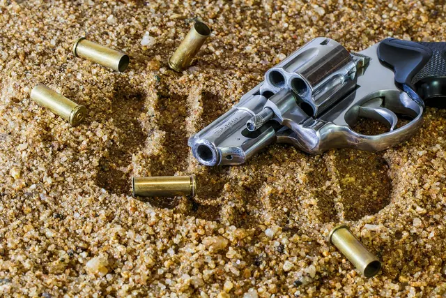 Stainless steel revolver with black grip lying in coarse sand, surrounded by scattered brass bullet casings