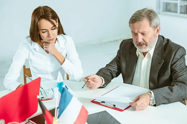 Two professionals at a white desk reviewing documents on a red clipboard, with small national flags in the foreground