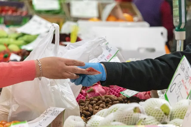 Customer hands cash to a gloved vendor at a market produce stall with peanuts, dragon fruit and handwritten price signs