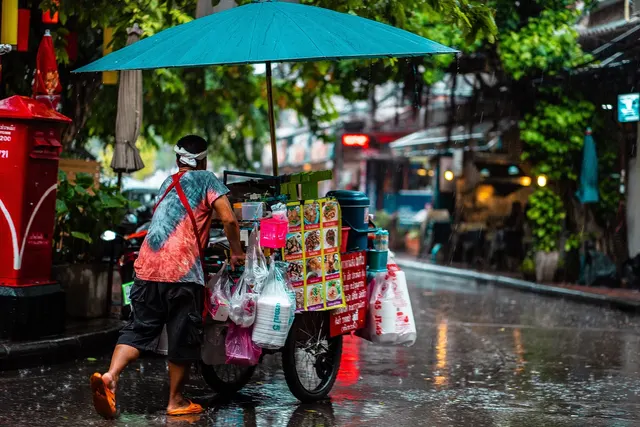 Street food vendor in a tie-dye shirt pushes a cart with a teal umbrella and picture menu along a rainy city street