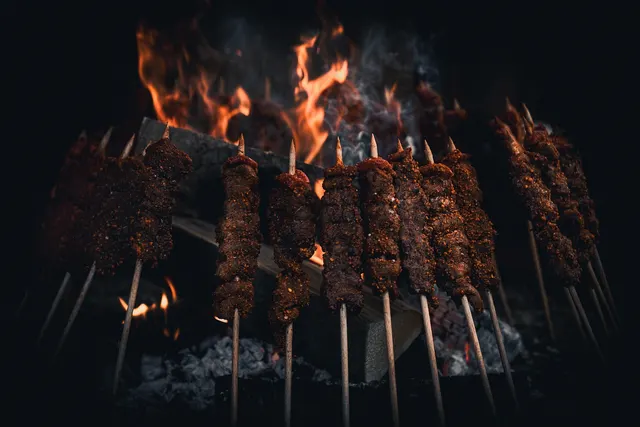Rows of seasoned meat kebabs on wooden skewers grilling over glowing charcoal and open flames against a dark background