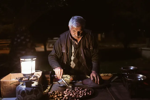Elderly street vendor roasting chestnuts on an outdoor cart at night, lit by a gas lantern beside a tray of nuts